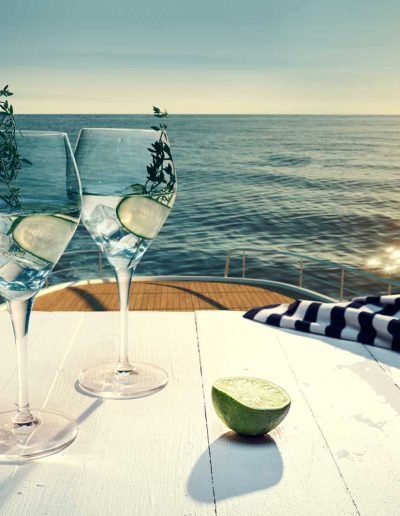 Clear beverage served in a wine glass, on top of a table that is on top level of a yacht, looking down onto the sea and horizon.
