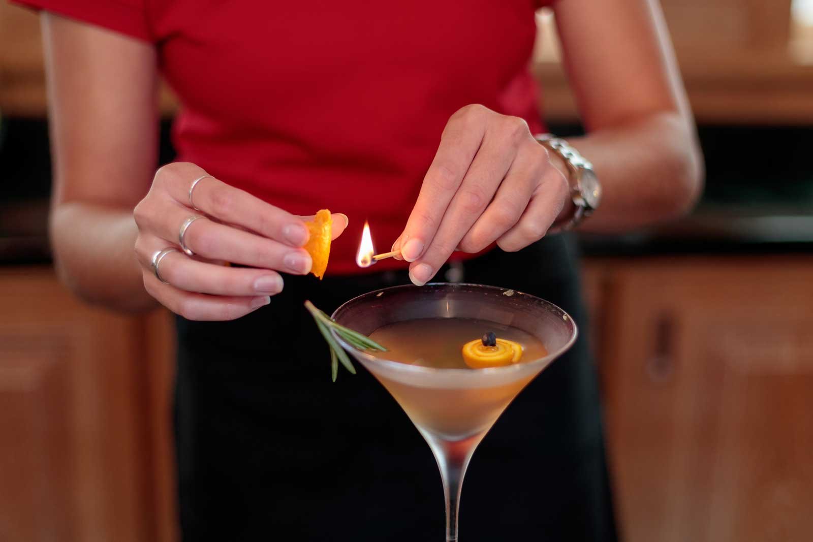 Woman squeezing an orange wedge to a flame of a match on top of a drink served in a martini glass with rosemary garnish and an orange wedge.