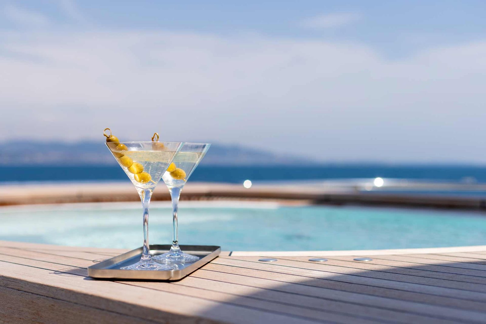 Two martinis with olives on a silver tray on the edge of a jacuzzi on the top deck of a yacht.