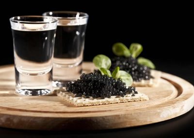 Caviar on a waffle cracker served on a wooden tray with two shot glasses of vodka.