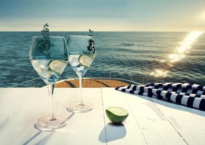Clear beverage served in a wine glass, on top of a table that is on top level of a yacht, looking down onto the sea and horizon.