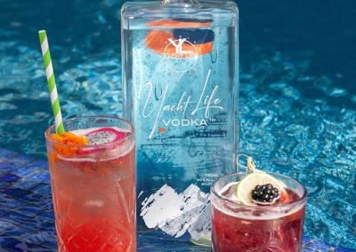 Yacht Life Vodka bottle on edge of a pool with two drinks in front of it.